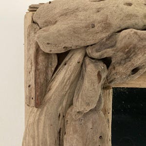 May include: A rustic mirror frame made from driftwood. The frame is made of natural wood with a rough texture and a dark brown color. The mirror is rectangular and has a black surface.