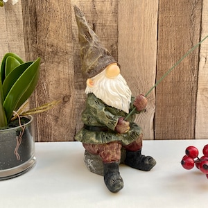 May include: A green and brown gnome figurine sitting on a rock, holding a fishing rod. The gnome has a white beard and a red hat. The gnome is sitting on a white surface.