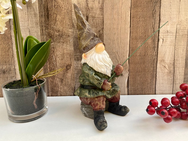 May include: A green and brown gnome figurine sitting on a rock, holding a fishing rod. The gnome has a white beard and a red hat. The gnome is sitting on a white surface.