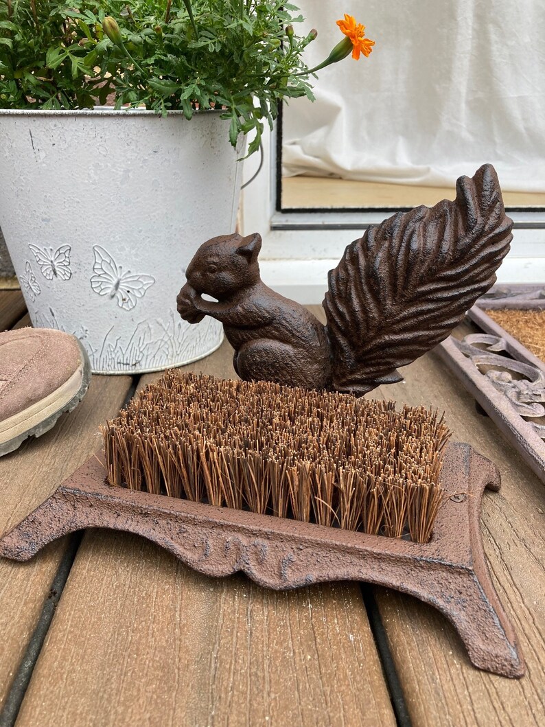 May include: A cast iron boot scraper with a squirrel design. The scraper has a brown finish and a stiff brush made of natural fibers.