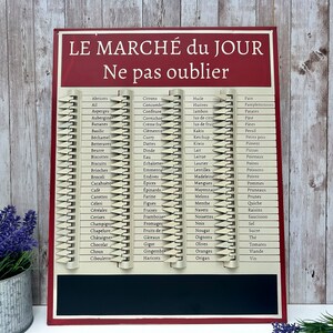 Op de afbeelding: Een rood en wit metalen bord met de tekst "LE MARCH&Eacute; du JOUR Ne pas oublier" in het Frans. Het bord heeft een krijtbord aan de onderkant en een lijst met boodschappen in het Frans aan de bovenkant.