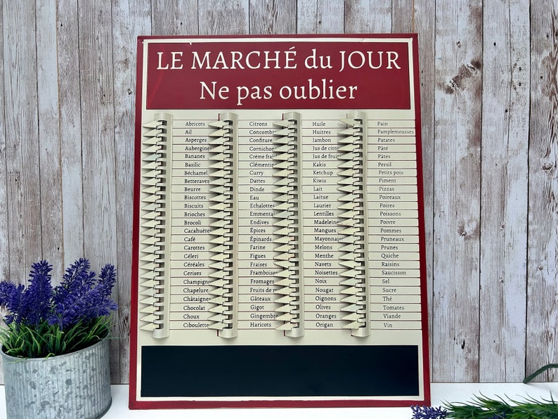 Op de afbeelding: Een rood en wit metalen bord met de tekst "LE MARCH&Eacute; du JOUR Ne pas oublier" in het Frans. Het bord heeft een krijtbord aan de onderkant en een lijst met boodschappen in het Frans aan de bovenkant.