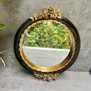 May include: Circular black and gold decorative mirror with floral detailing. The mirror features a black frame, a gold inner rim, and gold floral decorations at the top and bottom. A decorative item.
