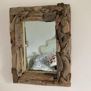 May include: A rectangular mirror with a unique frame made of driftwood. The frame is made of natural, weathered wood pieces that have been arranged in a decorative pattern. The mirror is reflecting a white bed with a patterned throw pillow.