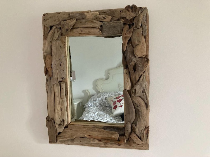 May include: A rectangular mirror with a unique frame made of driftwood. The frame is made of natural, weathered wood pieces that have been arranged in a decorative pattern. The mirror is reflecting a white bed with a patterned throw pillow.