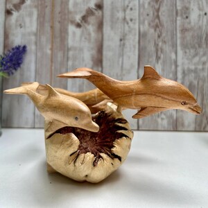 Peut inclure: Sculpture en bois repr&eacute;sentant trois dauphins sautant d'une base textur&eacute;e de couleur claire. Les dauphins sont sculpt&eacute;s dans un bois brun clair, avec des nageoires d&eacute;taill&eacute;es et des corps profil&eacute;s. La sculpture est plac&eacute;e sur un fond flou.