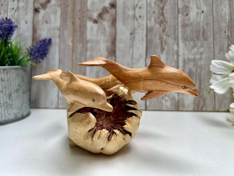 Peut inclure: Sculpture en bois repr&eacute;sentant trois dauphins sautant d'une base textur&eacute;e de couleur claire. Les dauphins sont sculpt&eacute;s dans un bois brun clair, avec des nageoires d&eacute;taill&eacute;es et des corps profil&eacute;s. La sculpture est plac&eacute;e sur un fond flou.