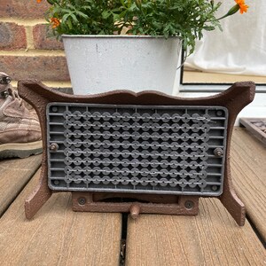May include: A brown cast iron fire grate with a black metal mesh insert. The grate is sitting on a wooden deck.