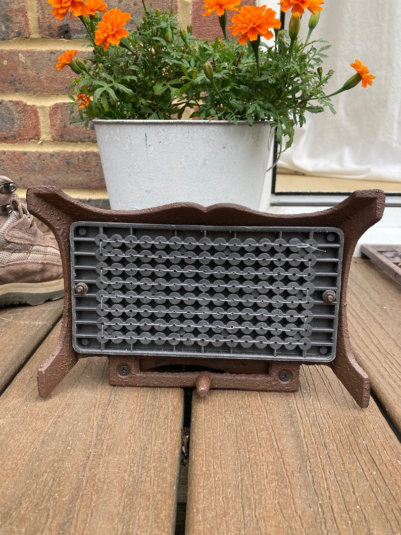 May include: A brown cast iron fire grate with a black metal mesh insert. The grate is sitting on a wooden deck.