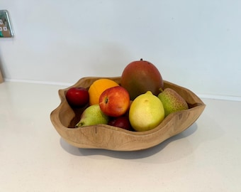 May include: A light brown wooden bowl with an organic, wavy edge shape containing a variety of fresh fruit, including a large mango, a bright yellow lemon, a green pear, a vibrant orange, and several small red apples of varying sizes.