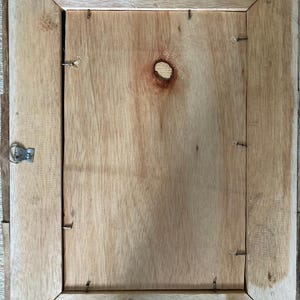 May include: The back of a wooden picture frame with a light brown finish. The frame has a simple design and is held together with small nails. There is a small hole in the center of the back panel.