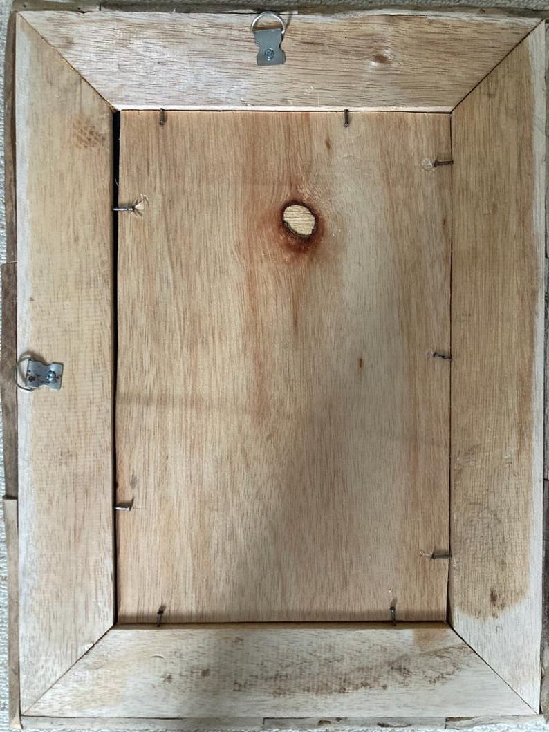 May include: The back of a wooden picture frame with a light brown finish. The frame has a simple design and is held together with small nails. There is a small hole in the center of the back panel.