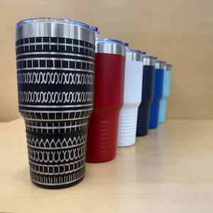 May include: A row of insulated tumblers in various colors, including black, red, white, navy blue, light blue, and a black tumbler with a silver geometric design. Each tumbler has a silver rim and a clear lid.
