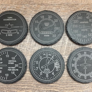 May include: Six black slate coasters with white printed aircraft instrument designs. The designs include a turn coordinator, RPM gauge, airspeed indicator, vertical speed indicator, attitude indicator, and compass.