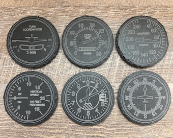 May include: Six black slate coasters with white printed airplane instrument designs. The designs include a turn coordinator, RPM gauge, airspeed indicator, vertical speed indicator, attitude indicator, and compass.