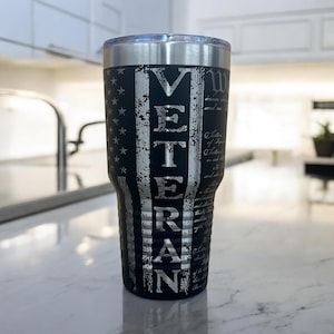 May include: Black insulated tumbler with a silver lid. The design features a distressed American flag with the word "VETERAN" in large, silver letters. The text of the Constitution is also visible on the side of the tumbler.