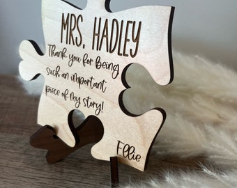 Pode incluir: Uma peça de quebra-cabeça de madeira com o texto "MRS. HADLEY Thank you for being such an important piece of my story! Ellie" gravado nela. A peça do quebra-cabeça está em pé sobre um suporte de madeira.