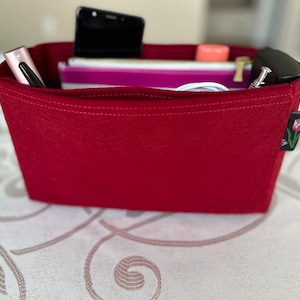 May include: A red felt bag organizer with a rectangular shape. The bag contains a black smartphone, a pen, a pink lipstick, a small zippered pouch, and other items. A small tag with a tulip design is attached to the side.