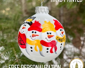 May include: Hand-painted Christmas ornament featuring three snowmen wearing red and yellow hats and scarves. The ornament is white with black eyes and buttons. The snowman on the right has the word "CHIEFS" written on its scarf.