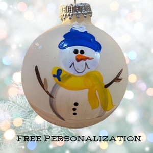May include: A white Christmas ornament with a hand-painted snowman wearing a blue hat and a yellow scarf. The snowman is holding a blue ribbon. FREE PERSONALIZATION text is visible at the bottom of the image.