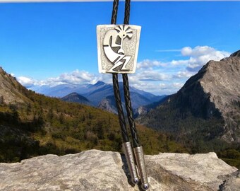 Op de afbeelding: Een zilveren bolo tie met een Native American Kokopelli-ontwerp. De stropdas is gefotografeerd tegen een bergachtige achtergrond.