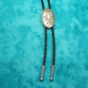 May include: Silver-toned bolo tie with an oval, engraved centerpiece. The centerpiece features a detailed geometric design. The black braided leather cord has silver-colored metal tips. The background is a teal fabric.