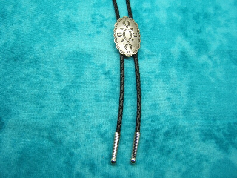May include: Silver-toned bolo tie with an oval, engraved centerpiece. The centerpiece features a detailed geometric design. The black braided leather cord has silver-colored metal tips. The background is a teal fabric.