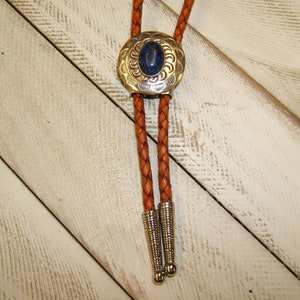 May include: A brown leather bolo tie with a silver and blue bolo tie slide. The slide has a decorative design and a blue stone in the center.