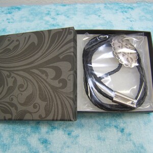 May include: A black leather bolo tie with a silver-colored decorative centerpiece, presented in a dark gray box with a swirling pattern. The tie is braided and has a silver-toned clasp.
