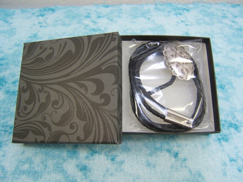 May include: A black leather bolo tie with a silver-colored decorative centerpiece, presented in a dark gray box with a swirling pattern. The tie is braided and has a silver-toned clasp.