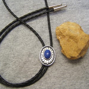 Puede incluir: Un corbatín de cuero negro con un colgante ovalado de plata y azul. El colgante presenta una piedra azul rodeada de un diseño floral plateado.