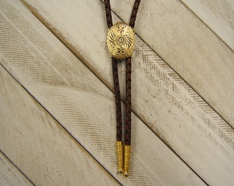 Könnte beinhalten: Ein braunes Leder-Bolo-Band mit einem ovalen Anhänger in Goldton, der ein Blumenmuster zeigt.