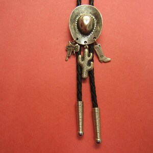 Puede incluir: Un bolo tie plateado con un dije de sombrero de vaquero, un dije de cactus y un dije de bota. La corbata es negra y tiene un cord&oacute;n trenzado.