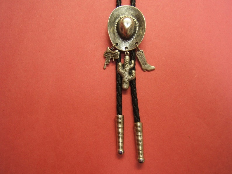 Puede incluir: Un bolo tie plateado con un dije de sombrero de vaquero, un dije de cactus y un dije de bota. La corbata es negra y tiene un cord&oacute;n trenzado.