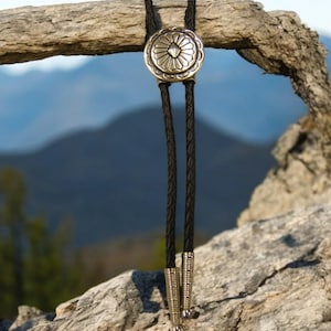 May include: A black bolo tie with a silver concho featuring a geometric design. The tie is hanging from a piece of driftwood with a blurred mountain background.
