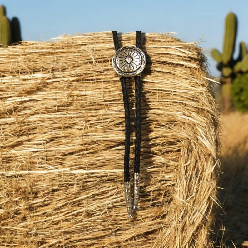May include: A black bolo tie with a silver concho featuring a geometric design. The tie is displayed against a background of a hay bale.