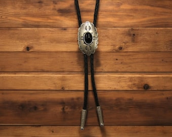 May include: A silver bolo tie with a black stone inlay and intricate designs. The tie is on a wooden background.