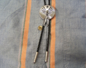 May include: A silver bolo tie with a black leather cord and a silver pendant featuring a Native American design. The pendant has a circular shape with a central design of a spiral and feathers. The bolo tie is worn on a blue and orange striped shirt.