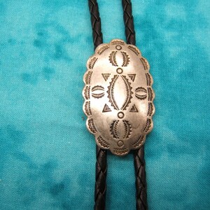 May include: Silver-toned bolo tie with an oval, patterned pendant. The pendant features geometric designs and is attached to a black braided leather cord. The background is a teal-colored fabric.