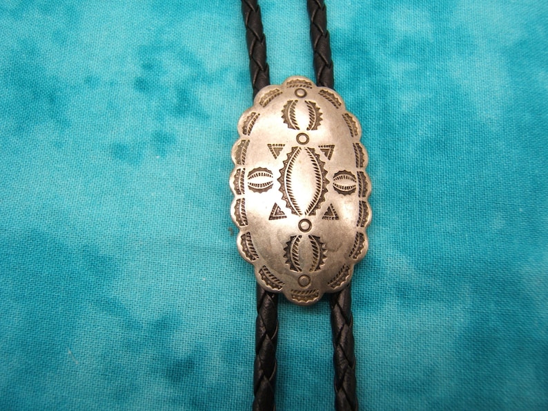 May include: Silver-toned bolo tie with an oval, patterned pendant. The pendant features geometric designs and is attached to a black braided leather cord. The background is a teal-colored fabric.