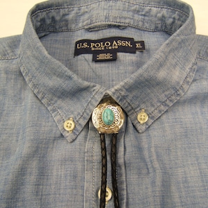 May include: A light blue button-down shirt with a U.S. Polo Assn. label and a bolo tie featuring a silver-toned metal setting with an oval turquoise stone. The bolo tie has a braided black cord and is fastened at the collar of the shirt.