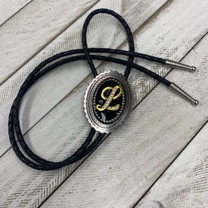Puede incluir: Una corbata bolo con un cordón trenzado negro y puntas de metal plateado. La pieza central ovalada tiene un borde plateado festoneado que rodea una incrustación negra con una letra L dorada adornada con pequeños cristales transparentes.