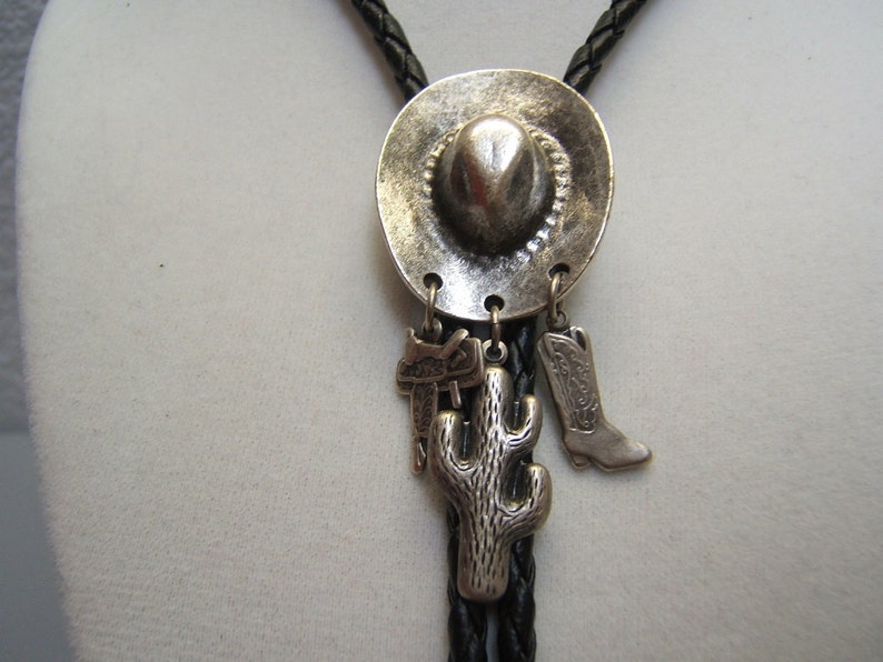 Puede incluir: Un bolo tie plateado con un dije de sombrero de vaquero, un dije de cactus y un dije de bota. El bolo tie est&aacute; hecho de cord&oacute;n de cuero negro.