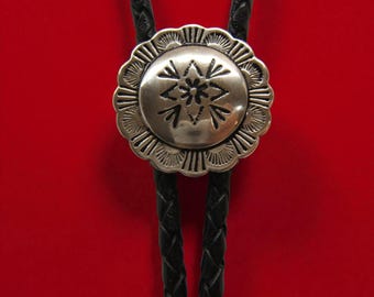 Könnte beinhalten: Eine silberne Bolo-Krawatte mit einem dekorativen Medaillon mit gewelltem Rand und einem geätzten Blumendesign. Das Medaillon ist an zwei schwarzen geflochtenen Lederkordeln vor rotem Hintergrund befestigt.