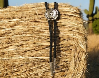 May include: A black bolo tie with a silver concho featuring a geometric design. The tie is displayed against a background of a hay bale.