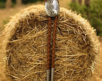 May include: A brown leather bolo tie with a silver concho featuring a floral design. The bolo tie is hanging on a bale of hay.