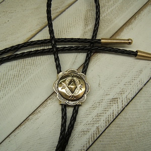 May include: A black braided bolo tie with a gold-toned, ornate, geometric design pendant.