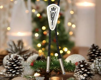 May include: A black bolo tie with a silver pendant featuring a geometric design. The bolo tie is displayed against a backdrop of a Christmas tree with lights and pine cones.