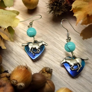 Puede incluir: Un par de pendientes de plata con una cuenta turquesa y un dije de caballo de plata en un colgante de corazón azul.