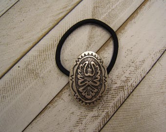 May include: Oval silver-tone hair tie with a detailed, embossed design. The decorative element features a central motif surrounded by intricate patterns and a scalloped edge. The black elastic band contrasts with the metallic centerpiece, creating a striking accessory.